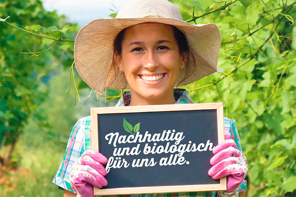 Eine lächelnde junge Frau mit braunem Haar und einem hellen Strohhut steht im Freien vor einem grünen Blätterhintergrund. Sie trägt Gartenhandschuhe und hält eine kleine Tafel mit der Aufschrift &quot;Nachhaltig und biologisch für uns alle.&quot; in weißer Kreide geschrieben. Die Frau trägt ein kariertes Hemd und strahlt Freude und Zufriedenheit aus, die bei den Österreichischen Mensen auf Regionaltität und Nachhaltigkeit hinweist.